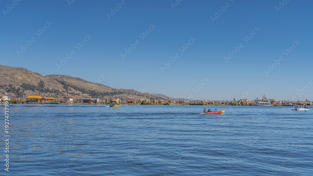 Fototapeta premium Tourist boats, reed boats balsa de totora sail on a calm alpine lake. Huts are visible on the shore of Uros Floating Island. Mountains against a clear blue sky. Peru. Puno. Lake Titicaca. Copy space