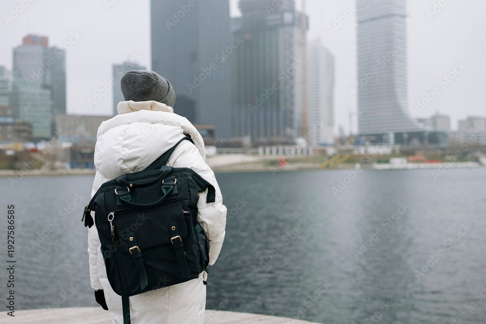 Obraz premium Young woman in white puffer jacket with a green backpack overlooks a foggy city waterfront. Concept of an urban winter activity