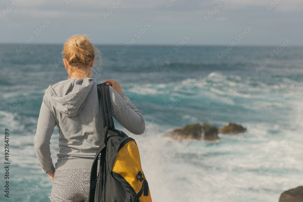 Fototapeta premium Young woman backpacker looking at ocean waves from rocky shore with copy space