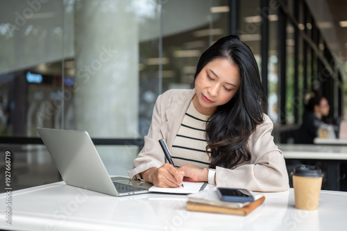 Asian woman student writing in notebook aside a laptop while sitting at table in university building