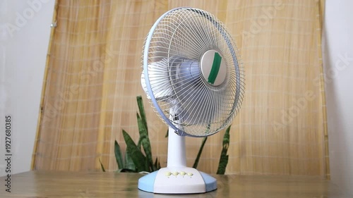 White and blue electric fan turns on, then the blades begin to spin, providing cooling relief from the heat