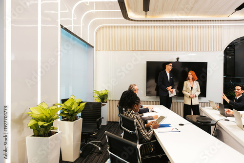 Tableau sur toile Modern conference room meeting with professionals collaborating around a long table, laptops open, two presenters by a screen, sleek lighting and indoor plants creating a contemporary office setting