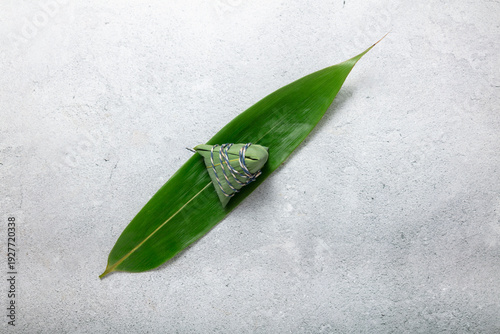 Fotografi Green chimaki dumpling wrapped in bamboo leaf resting on a larger leaf over gray