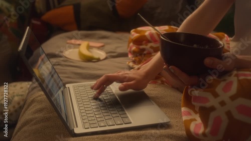 Close up shot of unrecognizable young woman using laptop and working from home while sitting on bed holding cereal bowl, focus on female hand typing, copy space