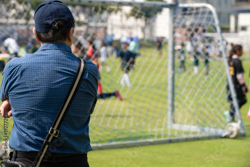 Candid back view of a supportive Asian father cheering for their son during a youth soccer match at school.