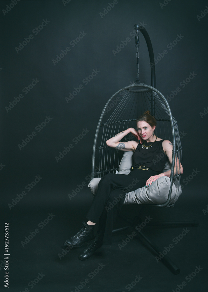 Fototapeta premium Ganzkörperportrait einer Frau im Hängesessel vor dunklem Studiohintergrund. Lässige Pose, moderne Ästhetik und stimmungsvolles Licht – Fashion trifft Interior-Design.