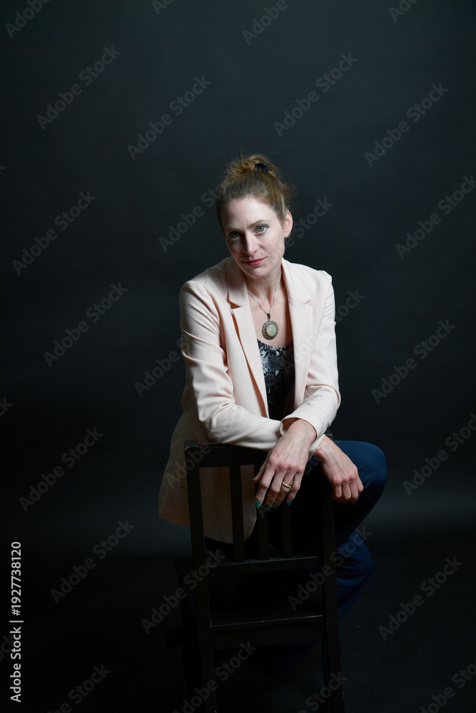 Fototapeta premium Stimmungsvolles Frauenportrait im Low-Key-Licht. Selbstbewusstes Model mit Blazer auf Stuhl vor dunklem Hintergrund – modernes Business- und Charakterportrait mit Tiefe.