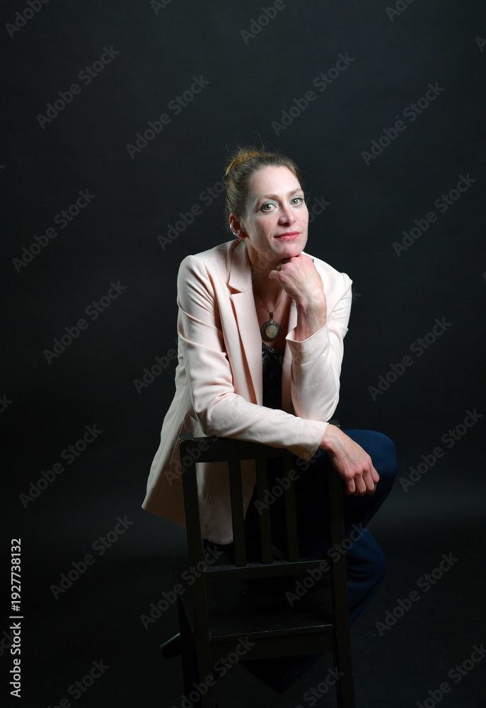 Fototapeta premium Stimmungsvolles Frauenportrait im Low-Key-Licht. Selbstbewusstes Model mit Blazer auf Stuhl vor dunklem Hintergrund – modernes Business- und Charakterportrait mit Tiefe.