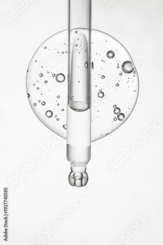 A glass cosmetology instrument with a transparent gel on a gray background, top view. A drop of serum and a pipette.