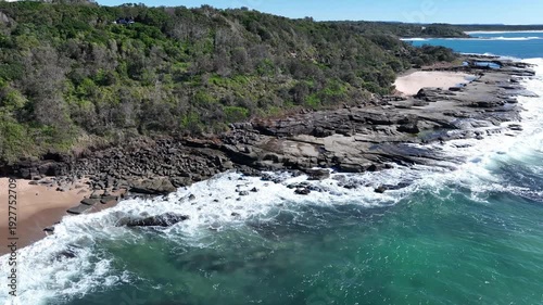 Blue water crashing against rugged rocks on a sunny day, white foam bursting into the air as bright blue skies shimmer and reflect the golden sun across the sparkling sea, Fast drone footage