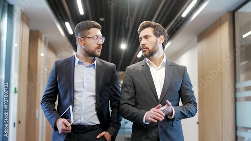 Two businessmen talking communicate while walking in the corridor of a modern business center. Colleagues in formal suits engaged in teamwork, discuss work issues. Professionals exchanging ideas