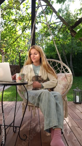 Middle aged woman working remotely with laptop and cat on her lap on terrace. Warm light, cozy lifestyle, family emotions, peaceful outdoor home office vibe.