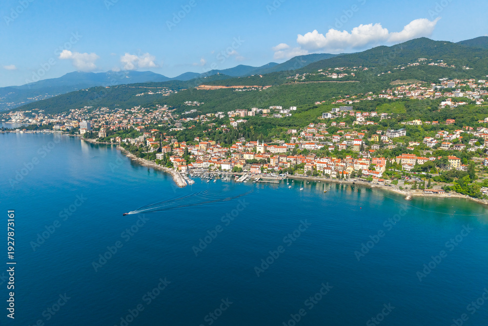 Obraz premium Aerial view of coastal town Volosko, Opatijska riviera, Croatia, with red rooftops along blue Adriatic Sea and green hills. Scenic Mediterranean landscape with marina and boat trail on calm water