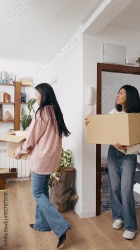 Young lesbian couple joyfully carrying cardboard boxes with their belongings, including a plant, while moving into their new home together. Vertical