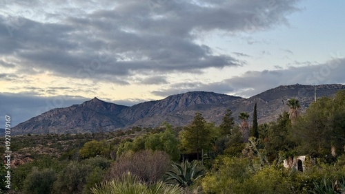 Serene Mountainscape: Underneath a dramatic, cloud-filled sky, a majestic mountain range rises, its rugged peaks and verdant foothills creating a breathtaking panorama. This awe-inspiring view.