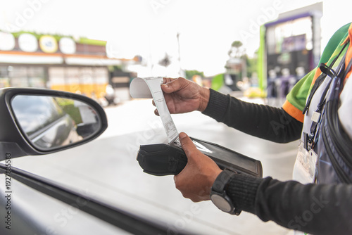 Close-up of a gas station worker holding a mobile payment terminal and pulling out a paper receipt for a customer at a petrol station pump.