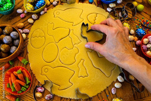 A hand carefully uses cookie cutters to cut out festive Easter cookies in the shapes of bunnies, eggs, and chicks from rolled out shortcrust dough.