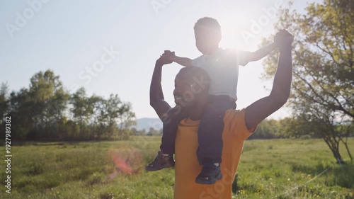 Cheerful father walking through a grassy field with his laughing son on his shoulders, both enjoying the sunshine and fresh air, slow motion shot.