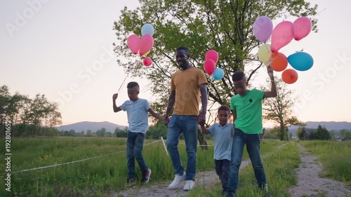 Happy African American family, a father and his boys walking with colorful balloons on a country path at sunset