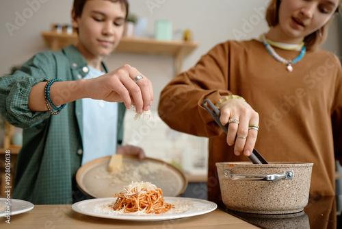 Tableau sur toile Teenage boy sprinkling grated cheese onto plate of spaghetti while teenage girl