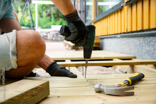 Homeowner using a power drill to install wooden deck planks during a backyard renovation, capturing realistic DIY, woodworking, outdoor construction.