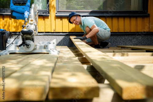 Man building wooden deck in backyard. Authentic DIY home improvement project during summer. Outdoor house renovation and construction.