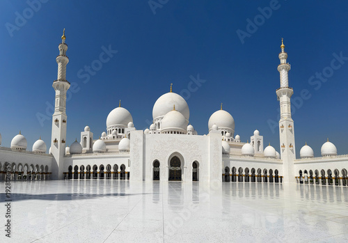 Panorama of Sheikh Zayed Grand Mosque, in Abu Dhabi, UAE. Pristine white marble domes and a tall minaret stand against a deep blue sky, perfectly reflected in the polished courtyard floor below.