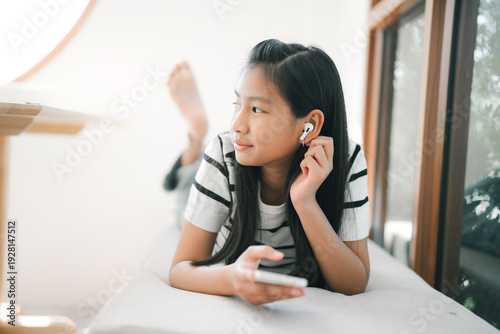 A young girl is lying on a sofa, holding a smartphone to her ear and smiling gently while looking at the camera in a bright, cozy room.