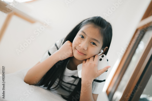 A young girl is lying on a sofa, holding a smartphone to her ear and smiling gently while looking at the camera in a bright, cozy room.