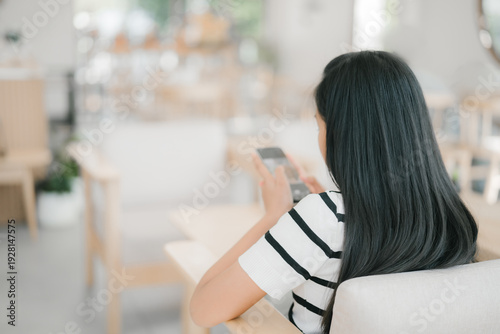 From behind, a young girl is sitting in a cozy café, playing on her smartphone and focusing on the screen while enjoying a quiet moment alone.