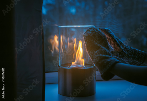 Obraz na plátne Small portable glass and metal bio fireplace burning bio ethanol gas liquid on living room window sill, child wearing winter gloves and warming hands indoors