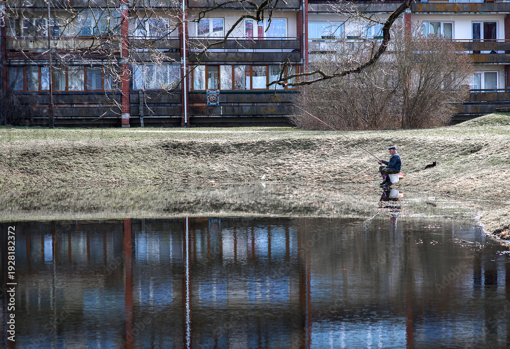 Naklejka premium Senior Man Fishing Alone By Urban Pond With Apartment Buildings Reflection