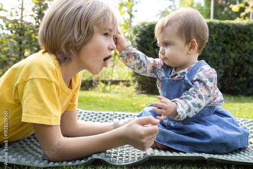 Two children, older brother younger sister, play together in garden, sitting on the grass. The little girl pulls the boy's hair, and he reacts emotionally. Summer walks, babysitting. Childhood moments