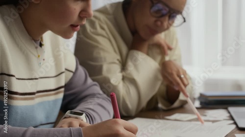 Close up of teen student writing on sheet of paper while her tutor guiding her and supporting