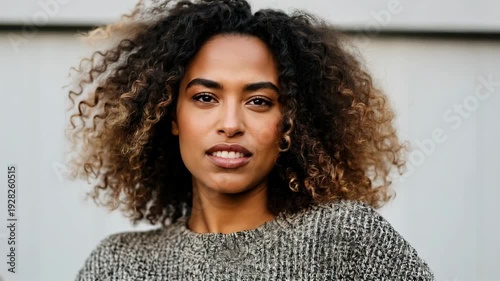 Smiling woman with curly hair wearing a gray sweater poses confidently in front of a light-colored wall, showcasing her joyful expression and engaging demeanor