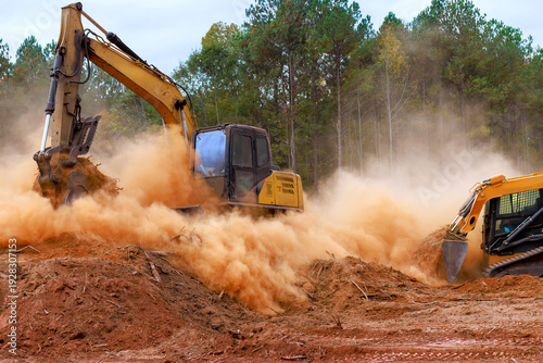 Construction skid steer equipment, excavator digs into ground, kicking up dust at site preparation land development.