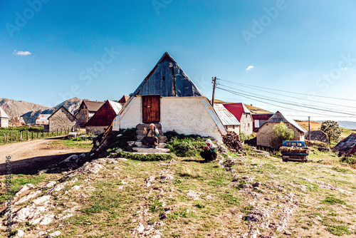 Lukomir, Bosnia and Herzegovina - 28 August 2015: View of an aged woman sitting in front of her traditional stone and wood house with a distinctive pyramidal roof, under a clear blue sky.
