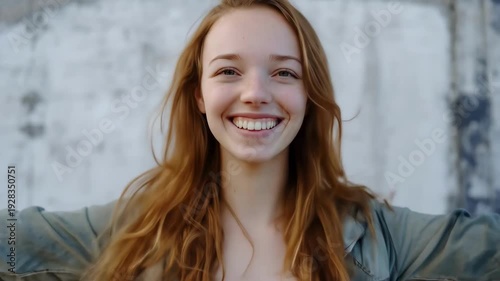 Smiling young adult Caucasian woman with long red hair and arms outstretched standing against a textured concrete wall.