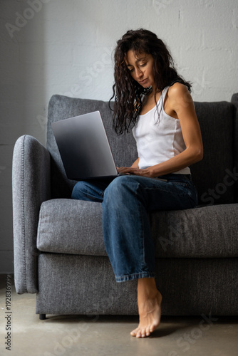 Free Download Concentrated young woman sitting on a sofa, working on a laptop with natural light coming from the window in a modern minimalist room. - GFXLoad