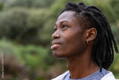 African American man gazing upward in park, showing gold hoop earring, dreadlocks, gray shirt