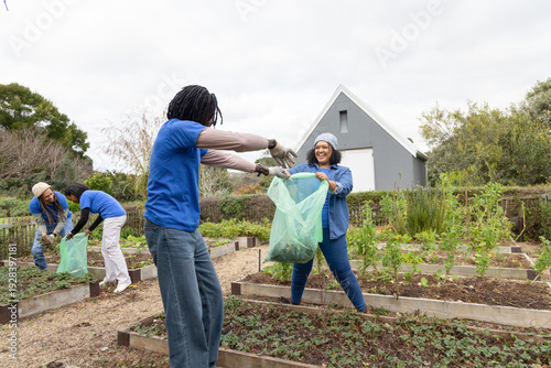 Diverse volunteers exchanging green compost bag in community garden by wooden beds wearing gloves
