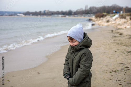 Tired six year old boy walking along beach in cold weather, child in winter jacket and hat looking down near sea shoreline