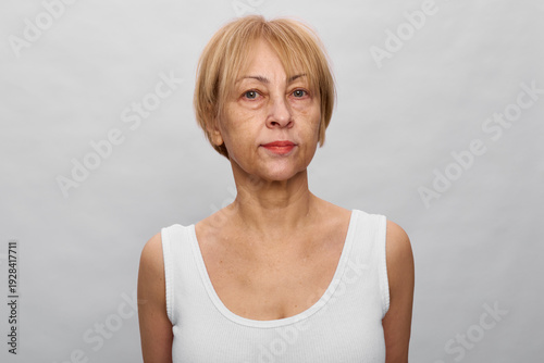 Bild auf Leinwand Portrait of a mature woman with short blonde hair wearing a white tank top, neutral expression on gray background