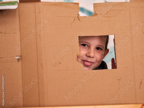Quadro su tela Smiling boy peeking through a hole in a cardboard box fort