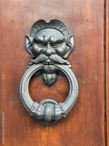 Florentine Devil Door Knocker on Wood - Ornate antique metal knocker with a menacing, grotesque devil or faun face and large ring, set against a warm, rustic wooden door in Florence, Italy.