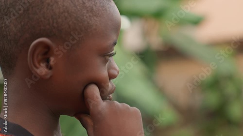 Bright young boy looks with gentle smile. Youthful boy with bashful expression among leafy trees in warm hues. Inquisitive young child gazes softly with shy smile amid lush greenery in warm colors