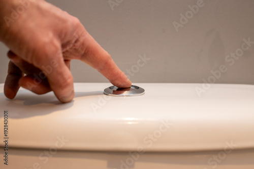 Close-up of a person’s hand pressing a chrome toilet flush button mounted on top of a cistern. Modern bathroom detail representing hygiene, sanitation, and everyday household routine.