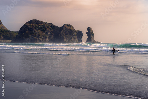 A surfer with a board walks into the ocean at Piha Beach, New Zealand, Auckland, with Lion Rock in the background, preparing to surf the waves on a cloudy day.