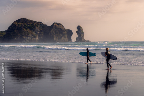 Obraz na plátne Two surfers in wetsuits walk along the beach with their surfboards after a surf session in Piha, Auckland, New Zealand, with Lion Rock in the background
