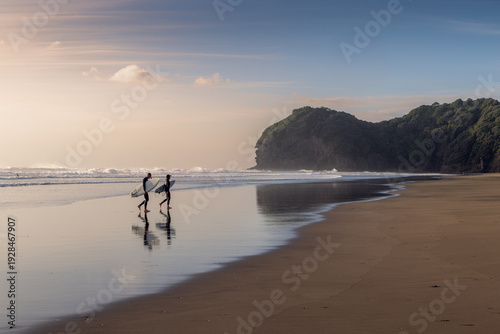Two surfers walk along the beach with their boards at Piha Beach in Auckland, New Zealand. They are walking towards Lion Rock after a morning surf session.